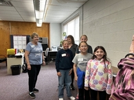 Inside a room with wood-paneled walls, a woman in a blue "Believe in Yourself" shirt stands with a group of four smiling 5th grade girls. The students are wearing name tags and casual school clothes.